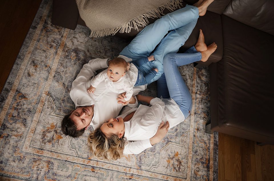 Ein Familienfoto mit einer jungen in weiß und jeansfarben gekleideten Familie bei einem Fotoshooting im Münchener Fotostudio. Bei dieser Aufnahme von oben guckt das kleine Baby der Familie in die Kamera.