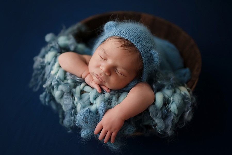 Süßes Neugeborenen Baby schläft in blauen Mütze mit Kuschlbär in einem Korb bei Babyshooting in München